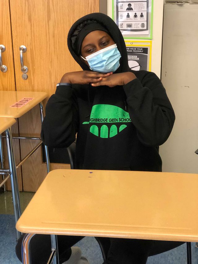 Fanta Sangare sits at a desk in a classroom; she is wearing a hooded sweatshirt and wears a medical masks, with her hands under her chin.